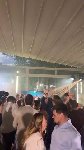 The video shows a crowded dance floor at an outdoor event. People are dancing and socializing under a white canopy ceiling. Lights and smoke effects are present, illuminating the scene. In the foreground, a man in a light blue shirt and beige pants is dancing with a woman wearing a white top. Behind them, a group of people, including women in dresses and men in shirts and suits, are also dancing and interacting. The background shows trees and a building with large windows, suggesting a venue with an open-air element. The overall atmosphere appears festive and energetic.