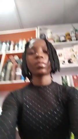 The video shows a young Black woman with braided hair, wearing a black shirt. She is standing in front of shelves stocked with various products, possibly in a store. The woman looks directly at the camera and then touches her hair, adjusting it on both sides of her head. The shelves behind her are filled with bottles, jars, and boxes.