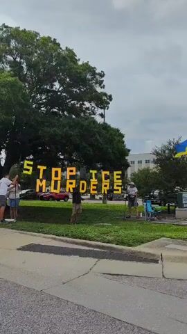 The video shows an outdoor scene. In the foreground, a sidewalk runs along the bottom of the frame, with a street to its left and a grassy area to its right. Several people are visible standing on the sidewalk. One man is holding a sign with yellow lettering, and a woman stands beside him. Further down the sidewalk, a person is seated in a wheelchair with a sign in front of them. A man is standing next to the person in the wheelchair. In the background, there are trees, cars, and buildings. The sky is overcast.