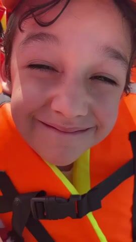 This is a close-up, first-person perspective video of a young child wearing an orange life jacket. The child is smiling widely, with their eyes crinkled. The camera is held very close to the child's face. In the first few seconds, a finger enters the frame and briefly touches the child's nose before being withdrawn. The child continues to smile throughout the video. The environment appears to be outdoors, possibly on a boat, given the life jacket and bright lighting.