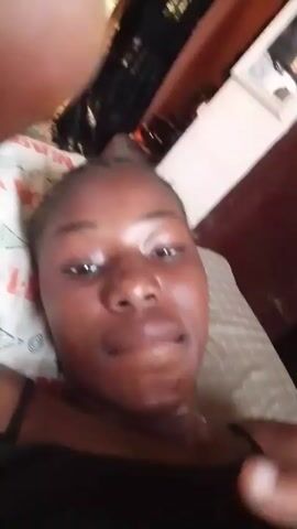 A dark-skinned woman is lying on a patterned pillow, facing the camera. She is wearing a black top. She smiles and sticks her tongue out slightly. Behind her is a white dresser with a mirror, a black curtain, and part of a brown wall. Background music is playing. A person's hand occasionally enters the frame from the top left.