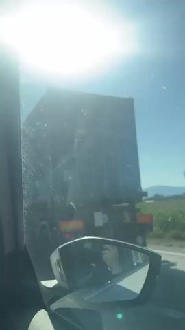 The video shows a view from inside a vehicle, looking out through the windshield and side mirror. The sun is very bright, causing lens flare and obscuring details of the sky. In the distance, there is a large truck or vehicle, possibly a garbage truck, moving on a road. The side mirror reflects a person's hand and part of the vehicle's interior. The environment appears to be outdoors, with trees and possibly hills visible in the background. The road seems to be a highway. The video is shaky, suggesting it was recorded while the vehicle was in motion.