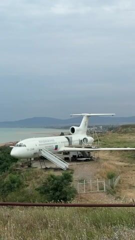 The video pans across a static airplane parked on a grassy hill overlooking the sea. The airplane is white with visible Russian text and a Russian flag on its fuselage. A set of stairs leads up to the plane's door. The background shows a body of water, a coastline with buildings and trees, and a hazy sky. In the foreground, there are dried grasses and wildflowers.