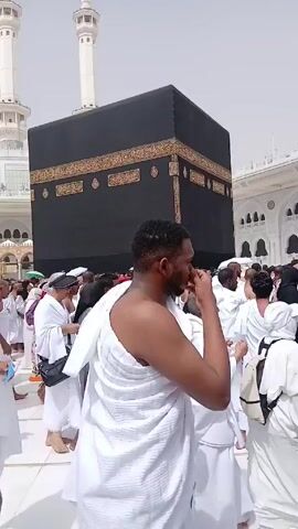 The video shows a large crowd of people in Mecca, Saudi Arabia. The pilgrims are dressed in white robes and are gathered around the Kaaba, a black, cube-shaped structure that is the most sacred site in Islam. The pilgrims are walking around the Kaaba. Some of them are praying, while others are simply observing the scene. There are two white minarets in the background.