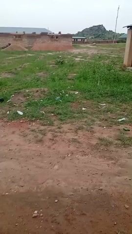 The video shows an outdoor scene with a dirt path in the foreground. To the left, a partially constructed brick building is visible, with piles of dirt and materials around it. The ground is covered with patchy green grass and scattered white plastic bottles and other debris. Further in the background, there are more buildings and a rocky hill. The sky is overcast and grey. The camera is moving slightly, giving a panning effect.