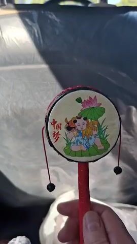 A hand holds a child's toy drum. The drum is red with black and red accents and has a round, flat surface decorated with an image of two children, bamboo stalks, and a lotus flower. Chinese characters are visible on the drum's surface, though they are not legible. A red string with a black bead is attached to the drum. The background is blurred, but appears to be the interior of a vehicle, with a dark, possibly leather-covered seat visible behind the drum. Sunlight casts shadows on the seat. The overall impression is that of a child's toy being played with or displayed within a car.