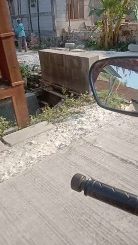 The video appears to be recorded from the perspective of someone riding a motorcycle or scooter, indicated by the presence of a rearview mirror in the upper left corner and a handlebar grip in the upper right. The footage is oriented vertically, suggesting the camera might be mounted on the vehicle. The environment seems to be a rural or semi-rural area under bright daylight, with a clear sky. The ground surface is a mix of concrete and gravel.

In the first few seconds, the scene is dominated by a light-colored, textured concrete surface and a bit of dry vegetation on the left. A pile of grey construction material, possibly cement or sand, is visible, along with some bricks and wooden scaffolding. In the background, there's a structure that looks like a building under construction, with white blocks and some wooden framing.

As the video progresses, the view shifts to a slightly different area. There are more piles of gravel and what appears to be dried material spread out on bamboo racks, suggesting agricultural activity, possibly drying tobacco or crops. A person wearing a blue and white striped shirt and a red cap is visible near these racks, seemingly engaged in some work. The construction continues to be present in the background. The video ends with a view of a concrete wall and green vegetation. The overall impression is a glimpse into a construction site and an agricultural area.