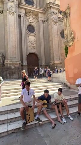 A group of young men are sitting on stone steps in front of a large, ornate building. One man is playing a guitar, another is playing a cajon drum. A third man is also sitting with them. In the background, several people are walking up and down the steps. The building appears to be a church or cathedral, with large wooden doors and intricate stone carvings. The sky is blue and the sun is shining.