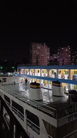 At night, a multi-story passenger ship is docked, with lights illuminated inside. The ship has white and blue paint, and two white funnels with gold and blue accents are visible on the top deck.  Several people are visible on the ship's decks, some standing and moving around. In the background, lighted buildings suggest a city skyline. The camera pans horizontally along the length of the boat. The audio contains sounds consistent with a ship’s engine or motor.