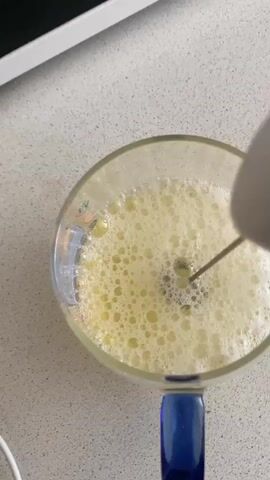 A close-up overhead shot shows a whisk attachment from a handheld mixer being used to froth a pale yellow liquid in a clear glass mug with a blue handle. The liquid is already bubbly and appears to be egg whites or a similar frothy substance. The mixer is held at an angle, and the whisk is submerged and rotating, creating more foam and splashes within the mug. The mug is placed on a speckled, light-colored countertop. In the background, the edge of a white appliance is visible, likely a counter or appliance.
