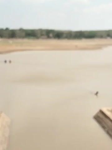 A person is fishing in a body of water. The person is in a small boat and holding a fishing rod. The water is a murky brown color. In the background, there is a dry, sandy shore with some trees and buildings. There are also a few other people wading in the water in the distance.