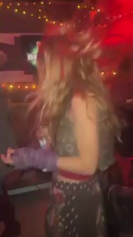 A woman is dancing at a party. She has long curly hair and is wearing a tank top, long skirt, fingerless gloves, and glasses. She is holding a water bottle in her left hand. The room is lit with red and green lights and decorated with string lights and balloons. Other people are visible in the background.