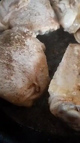 The video shows a close-up view of several pieces of chicken cooking in a pan. The chicken pieces appear to be bone-in thighs or drumsticks, seasoned with spices, and are in the process of browning. The pan appears to be dark-colored and is likely non-stick. There is a slight shimmer of oil or rendered chicken fat in the pan, indicating that the chicken is actively cooking. The lighting is somewhat dim, but the focus is on the chicken.