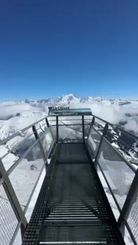 The video shows a viewpoint at the La Clusaz ski resort in the French Alps. The camera is positioned on a metal walkway with a metal railing on either side, looking out towards a panoramic view of snow-covered mountains. There is a sign that says "La Clusaz" above a picture of the resort. The sky is a clear, bright blue, and there are clouds nestled between the peaks of the mountains. In the distance, a taller, snow-capped mountain stands out above the clouds.  Someone is heard speaking in the background, but they are not visible. The overall impression is of a clear, sunny day at a high-altitude vantage point.