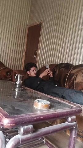 A young man is seen lying on a brown sofa, dressed in a black shalwar kameez. He is holding a smartphone with both hands and appears to be intently looking at the screen. In front of him, there's a glass-top table on which a silver pitcher and an ashtray are placed. The background features a wall with vertical beige paneling and a dark brown door. The scene takes place indoors, likely a living room or lounge area, in Samundri, Pakistan. The man's expression is neutral as he engages with his phone. The video includes a brief zoom-in on the man, followed by a quick, blurred pan of the scene.