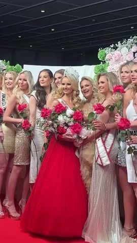 The video shows a group of women in various dresses, posing on a red carpet. Most of them are wearing short, shiny dresses in gold or silver and holding a single red rose.  Several are wearing sashes with their country’s name. At the center of the group, a woman wearing a tiara and a large red gown holds a bouquet of pink flowers. The background features a wall decorated with white and pink flowers and curtains. The scene is indoors, under a high ceiling with visible lighting and rigging. The camera zooms out, showing the whole group and the size of the room.