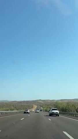 The video shows a view from inside a moving vehicle on a highway. The sky is clear and blue. The highway is asphalt with white lane markings. There are several other vehicles on the road, including a white SUV in the right lane, a white sedan in the left lane, and several other cars further ahead. The landscape on either side of the highway appears to be desert with low-lying brush and hills in the distance. There are metal guardrails along the sides of the road. A green highway sign is visible on the right side of the road in the distance.