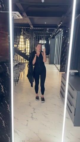 A woman stands in front of a mirror, taking a selfie. She is wearing a black jumpsuit with a zipper down the front and black sneakers. She is also carrying a black jacket over her left shoulder. Her hair is blonde and pulled back. She has tattoos on her left arm and right wrist. The background appears to be a modern interior with a staircase and black marble accents. The lighting is bright, with vertical light fixtures on either side of the mirror.