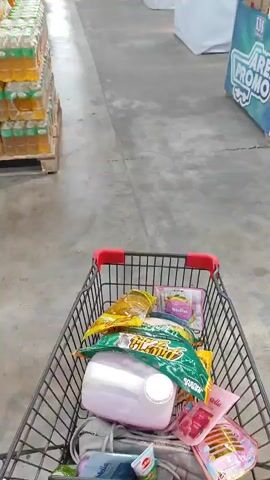A first-person perspective video shows a shopping cart filled with various items, including drinks, snacks, and a large white plastic container. The cart is being pushed down an aisle in a store. To the left, shelves are stocked with rows of bottled drinks. A young girl in a pink shirt enters the frame from the right and briefly interacts with the shopping cart.