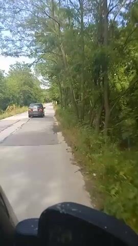 The video shows a first-person perspective of someone riding a vehicle, possibly a motorcycle or ATV, down a paved road. The road is flanked by trees and vegetation on both sides. There is a black car ahead on the road, and a road sign on the right side of the road. The camera is mounted low, likely on the vehicle's handlebars or front, and some of the vehicle's parts are visible in the foreground. The sky is partly cloudy and the lighting suggests it is daytime. The rider appears to be moving forward at a moderate pace.