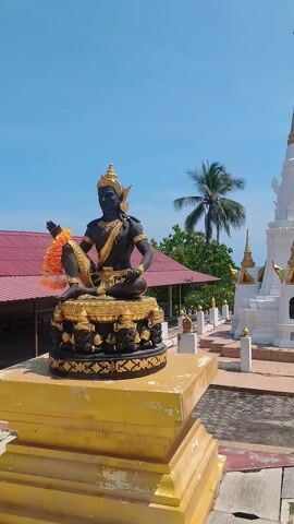 The video shows a temple complex in Thailand on a sunny day.  The first shot is of a black and gold statue of a deity.  The next shots show a white pagoda with a gold spire, a pavilion with a red roof, and a view of the ocean. There is a person walking near the pavilion.