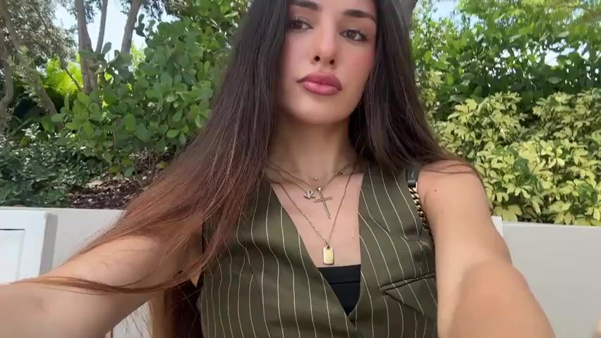 A woman with long, dark hair and makeup is taking a selfie outdoors. She is wearing a green pinstripe vest over a black top and several necklaces, including a cross pendant and a letter pendant. The background shows greenery and a white structure. She is looking at the camera and puckering her lips.