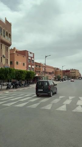 The video shows a street scene in a Moroccan city.  The buildings are mostly multi-story and made of beige and reddish-brown materials. Some have decorative architectural features. There is a crosswalk with white stripes on the street. A dark van drives across the crosswalk. The street is lined with trees. There is a cafe named "Cafe Hema Restaurant" with outdoor seating where people are sitting.  Next to the cafe is a store named "Beqalaty." A man walks in front of the store, looking at his phone.  The sky is overcast.