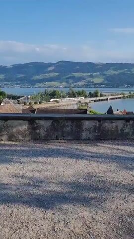 A person is standing on a gravel path, looking out at a scenic view of a lake and hills. The sun is shining, and there are some clouds in the sky. A stone wall runs horizontally across the foreground, partially obscuring the view of buildings and a body of water in the middle ground. A bridge crosses the water in the distance, and rolling hills covered in greenery form the background.