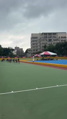 A group of young people on roller skates are racing on a green track in front of a building. The sky is overcast. There are tents in the background.