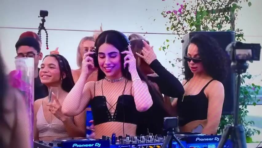 The video shows a DJ playing music at an outdoor party, surrounded by people. The DJ is a young woman wearing a black top, headphones, and a silver chain necklace, and is operating a Pioneer DJ controller. Several people are standing around the DJ, some holding drinks. A person on the right wears sunglasses. There is a speaker, a camera on a tripod, and plant foliage visible in the background. The overall environment suggests a lively and celebratory atmosphere.