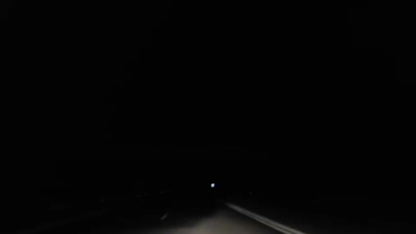A first-person perspective from a vehicle shows a dark road at night. The vehicle's headlights illuminate the asphalt and double white lines in the foreground. Distant red lights are visible on both sides of the road. Periodically, flashes of lightning briefly illuminate the dark sky and distant landscape. A white road sign is visible on the right side of the road.