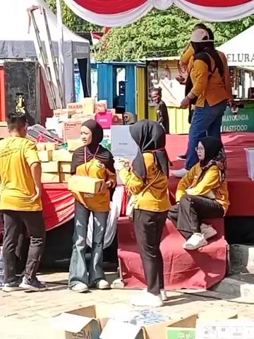 The video shows a group of people, most of whom are wearing yellow t-shirts with a logo and some are wearing headscarves. They appear to be at an outdoor event, possibly a market or festival, with tents and stalls in the background. Some individuals are holding boxes, suggesting they are distributing or selling items. One person is speaking into a microphone. The overall atmosphere seems to be one of activity and organization.
