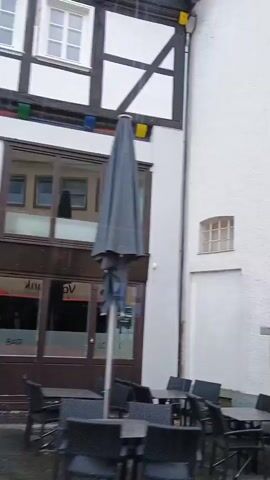 The video shows an outdoor view of a building with white textured walls and a large pointed archway. To the left of the archway, there is a building with a dark timber-framed upper story and a white lower story. A small, barred window is visible on the white wall to the left of the arch. Tables and chairs are set up outside the building to the left. The camera pans to the right, showing the archway and a glimpse of an indoor space through a glass window, where stacked chairs and a table are visible. The view then shifts to an outdoor seating area with people sitting at tables under an umbrella. A person wearing a dark jacket is standing and appears to be interacting with the seated individuals. The scene suggests a cafe or restaurant setting in an urban environment, possibly in Europe due to the architecture.