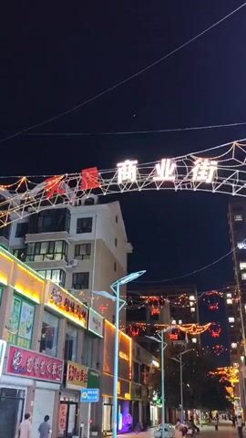 The video shows a street scene at night, illuminated by decorative lights. On an archway spanning the street, there are Chinese characters and strings of lights. Buildings lining the street are lit up with colorful lights, some displaying signs with Chinese characters. There are streetlights and more strings of lights with red lantern decorations hanging across the street. Pedestrians are visible walking on the sidewalk, and a person is riding a scooter. A woman is standing in the foreground, looking up. The ground has circular light projections.