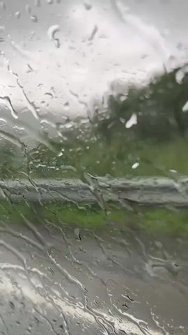 The video is filmed through a wet window, possibly a car windshield, as raindrops distort the view of the outside environment. The outside scene is blurry and shows a road with some buildings and greenery. In the background, there is a cell tower. There are no people or specific actions visible, only the visual effect of rain on glass.