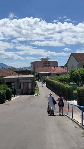 The video shows a street scene with a few people and buildings. Two individuals, one wearing a white robe and the other in athletic wear, are walking down a paved road. Behind them, another person is walking away from the camera further up the road. The environment is characterized by houses with red tiled roofs, green hedges, and a blue sky with scattered white clouds. In the background, a large, light-colored building with multiple floors is visible. The overall impression is of a sunny day in a residential area.