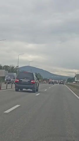 A point-of-view shot from a vehicle traveling on a highway. Several cars and trucks are visible on the road, moving in the same direction. The sky is overcast, with a mountain range visible in the distance. The highway has multiple lanes, separated by white lines. There are streetlights and red-and-white traffic barriers along the side of the road. The overall atmosphere is one of normal highway traffic under a cloudy sky.