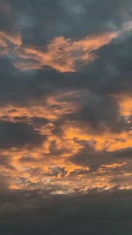 A video shows a close-up view of dramatic sunset clouds. The clouds are a mix of dark gray and vibrant orange, with the orange light illuminating the undersides of the clouds and creating a striking contrast. The camera is stationary, capturing the stillness of the sky.
