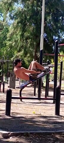 A man is doing crunches on a decline ab bench at an outdoor park. The man is shirtless and wears sneakers, shorts, and possibly underwear. The decline bench is black and silver and is located next to a series of black exercise bars. The ground is covered with loose dirt, and a few trees are visible.