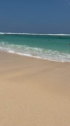 The video captures a serene and picturesque beach scene on a bright, sunny day. The sky above is a clear, deep blue, devoid of clouds. The ocean water is a striking turquoise green, with white-crested waves breaking in the distance, indicating a calm but active sea. Closer to the shore, the water appears clearer, allowing a glimpse of the sandy bottom through its light turquoise tint.

In the foreground, a stretch of pristine, light beige sand is visible. A gentle wave with white foam rolls onto the shore, momentarily washing over the sand before receding, leaving behind a thin, shimmering layer of water. There are a few individuals visible in the ocean, swimming casually in the clear, inviting water. One person is seen swimming in the mid-distance, and another appears further out. The scene evokes a sense of tranquility and natural beauty, highlighting the clear waters and soft sands typical of a tropical or idyllic beach destination.