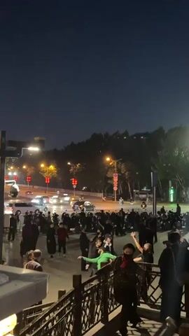 At night, a group of mostly older people are dancing in a public space near an intersection with traffic. The group is standing on a paved area adjacent to a railing. They are clustered together, some in pairs, dancing to music. Behind them, there's a street with moving cars, traffic lights, and parked motorcycles. There are trees and streetlights visible, creating a dimly lit atmosphere. The camera is positioned from a slightly elevated angle, looking down at the dancing crowd.