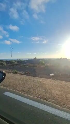A side view from a car shows a sunny coastal landscape. The sky is blue with scattered white clouds. The sun is visible on the right side of the frame, shining brightly. In the middle ground, there is a dry, rocky terrain with sparse vegetation and a tall, spiky plant. The ocean is visible in the distance, meeting the horizon under the bright sky. The foreground shows a paved road with a white line on the left edge and the side mirror and window frame of the car. The camera appears to be moving, suggesting the video is a travelogue.