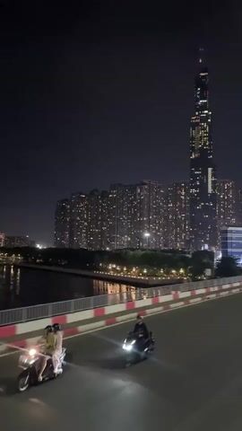 This media file captures a night scene in Thủ Đức, Vietnam, featuring a modern cityscape dominated by a very tall skyscraper and a cluster of large apartment buildings. The time of day is clearly night, with the sky being a deep, dark blue, hinting at a clear night with minimal cloud cover. The buildings are illuminated with numerous lights, creating a warm glow against the darkness and reflecting on the water body in the mid-ground.

In the foreground, a bridge is visible with a road that has a pink and white striped barrier along the side. Two people are riding on a scooter on this bridge. The scooter's headlights are on, cutting through the night. The rider and the passenger are both wearing helmets, indicating safety consciousness. Their presence on the bridge suggests they are commuting or sightseeing. The atmosphere appears to be calm and urban, with the city lights creating a sense of vibrancy.

The prominent skyscraper, likely Landmark 81, is a striking feature of the skyline, its height and illuminated windows emphasizing its architectural significance. The surrounding apartment buildings suggest a densely populated urban area. The presence of a body of water, possibly a river, adds a reflective element to the scene, further enhancing the visual appeal of the city lights. The overall impression is one of a bustling, modern city at night, captured from a vantage point on a bridge. No visible text is present in the media file.