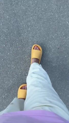 The video shows a first-person perspective of someone walking on a grey asphalt surface. They are wearing white pants with red stains and yellow slide sandals. To their right, another person wearing black slide sandals is walking alongside them. The scene is outdoors.