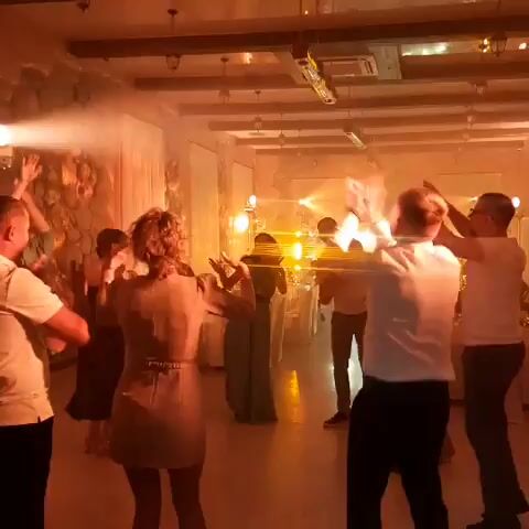 The video shows a group of people dancing in a dimly lit room. The room appears to be a banquet hall or event space, with stone walls and a wooden ceiling. There are tables and chairs set up in the background, and string lights are hanging from the ceiling. The people are dressed in formal attire, and they are all smiling and having a good time. The video is shot from a low angle, and it is slightly shaky. The lighting in the room is warm and inviting, and there is a haze of smoke or fog in the air.