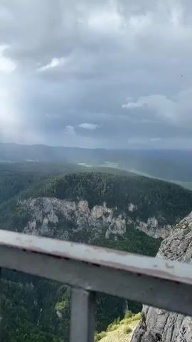 The camera pans upwards to show a view of a mountain range under a cloudy sky with patches of sunlight. The foreground shows a metal railing, slightly rusted. The mountains are covered in dense, green trees, with exposed rock faces in some areas. The sunlight streams down in visible shafts through the clouds.
