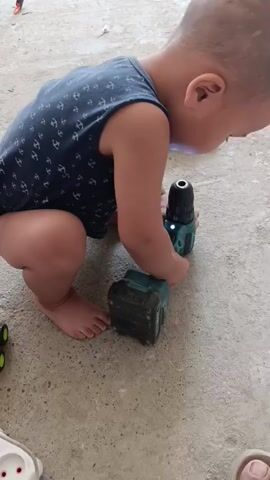 A toddler wearing a dark blue sleeveless shirt with a pattern of white anchors is crouched on a concrete surface. The toddler is interacting with a teal and black cordless drill and a teal battery pack. The drill is positioned on top of the battery pack. The toddler's right hand is touching the drill, and their left hand is near the battery pack. The toddler's legs are bent, and their bare feet are visible. The environment appears to be outdoors or in an open, unpaved area, with concrete flooring and some lighter colored gravel or dirt in the background. The lighting suggests it is daytime.