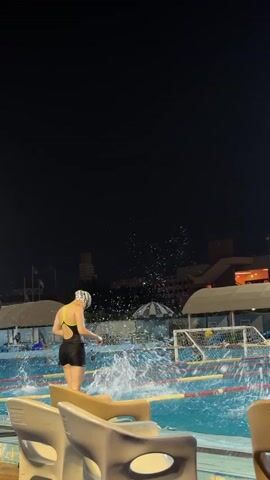A person in a black and yellow swimsuit and a white swim cap stands at the edge of a swimming pool at night. They are facing away from the camera and towards the pool. In the background, several other people are swimming in lanes marked by yellow and red ropes. A water polo goal is visible in the right side of the frame. The person then dives into the pool.