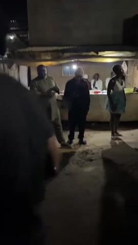 At night, under a makeshift shelter, a group of people are gathered. In the foreground, a man in a light-colored outfit and a man in a dark outfit are facing each other and appear to be interacting. Behind them, at a counter, several individuals are visible. One person is wearing a white hat, and two others appear to be seated. To the right of the counter, a woman in a light-colored dress stands facing away from the camera. The ground is uneven and appears to be dirt. Shadows are cast on the ground, suggesting artificial lighting.