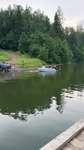 A dark-colored SUV is parked on a dirt ramp at the edge of a body of water, with its rear facing the water. A trailer is attached to the SUV, and a light-colored motorboat is partially submerged in the water, connected to the trailer by a blue strap or rope. A person is visible in the boat, sitting in the driver's seat and appearing to operate the motor. The boat is positioned in the water as if it is being launched or retrieved. The surrounding environment is natural, with lush green trees and vegetation on the bank behind the SUV and extending into the background. The sky appears overcast, suggesting cloudy weather. In the foreground, the edge of a wooden structure, possibly a dock or pier, is visible, with part of a person's back and shoulder wearing a hooded jacket facing away from the camera. The water is calm and reflects the trees and sky.